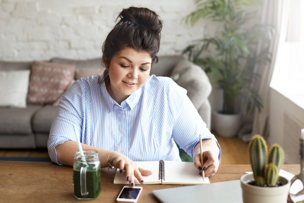 Hardworking positive young female freelancer with heavy curvy body multitasking at home office, texting sms on blank screen mobile phone and making notes in her diary, writing down plans and ideas